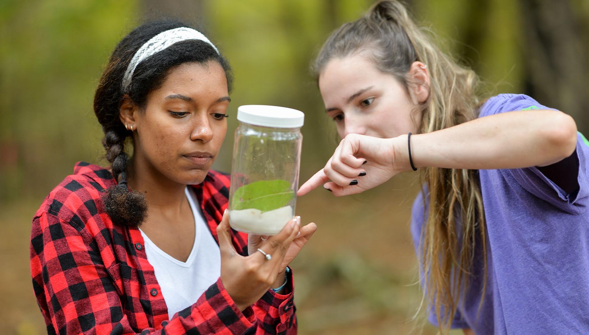 two female students examening plants and specimen in a glass jar